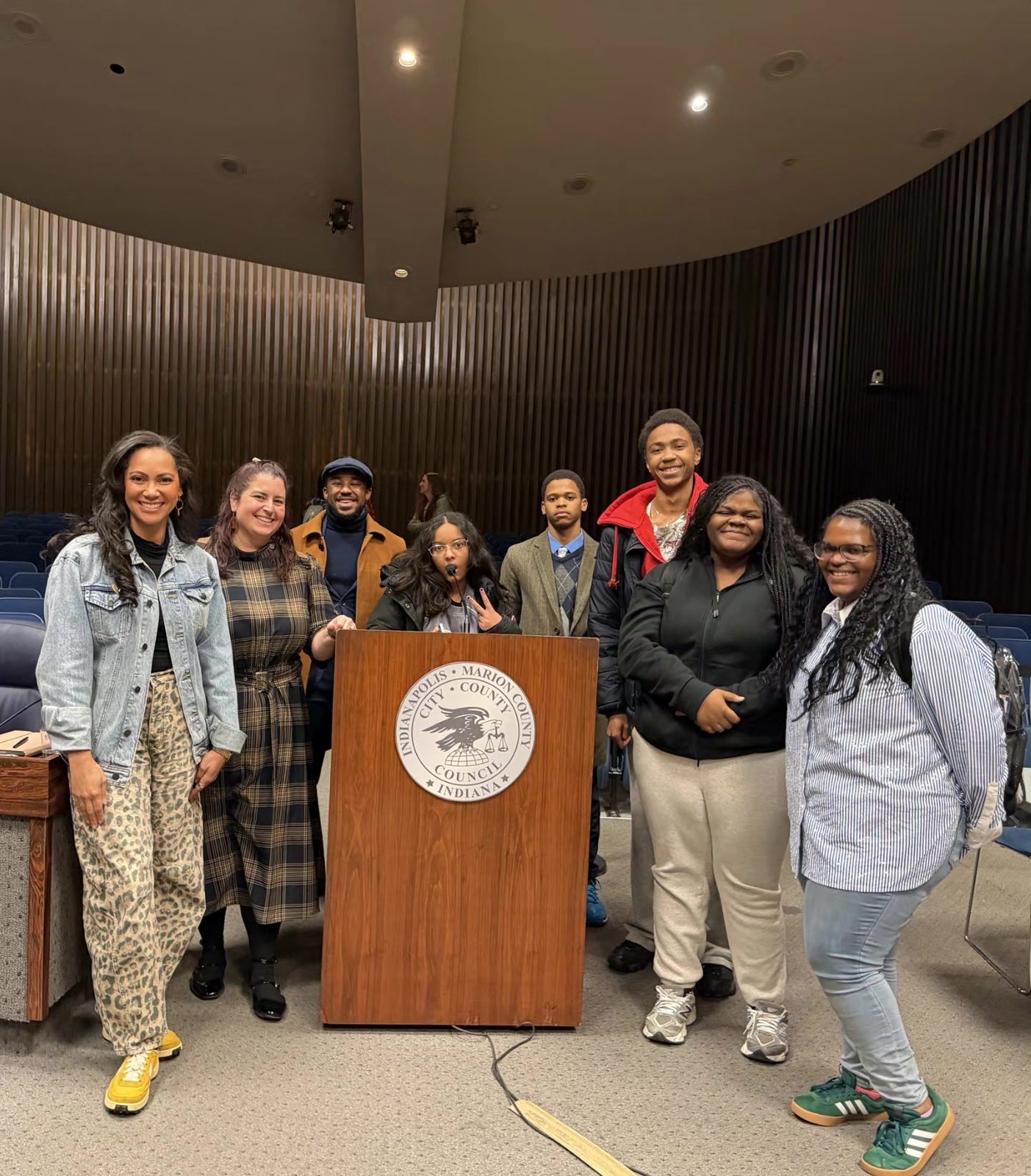 Students and adults smile around the podium in the Indianapolis City-County Council chambers