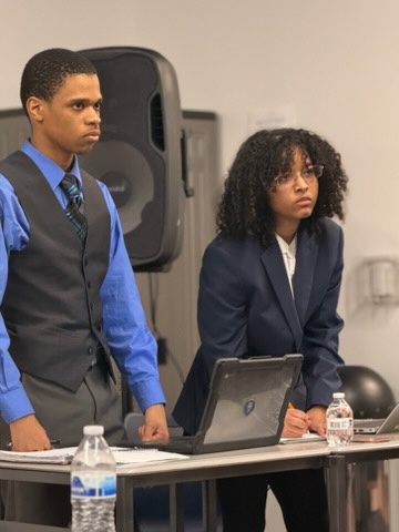 Two students stand at a table with laptops, listening during a debate round
