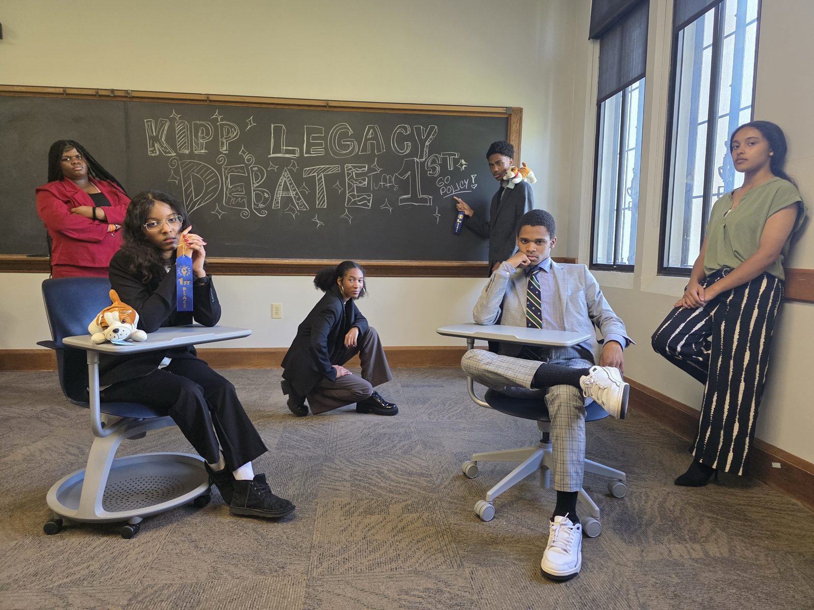 Debate team members pose in a classroom with first-place ribbons in front of a chalkboard reading KIPP Legacy Debate 1st