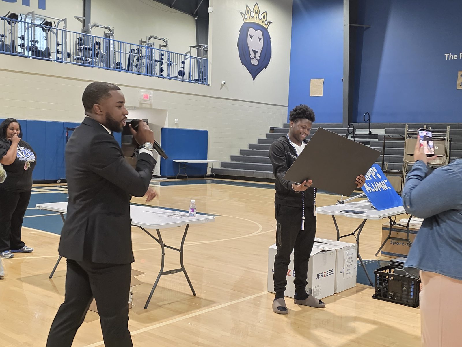 The presenter speaks into a microphone as the graduate looks at the check