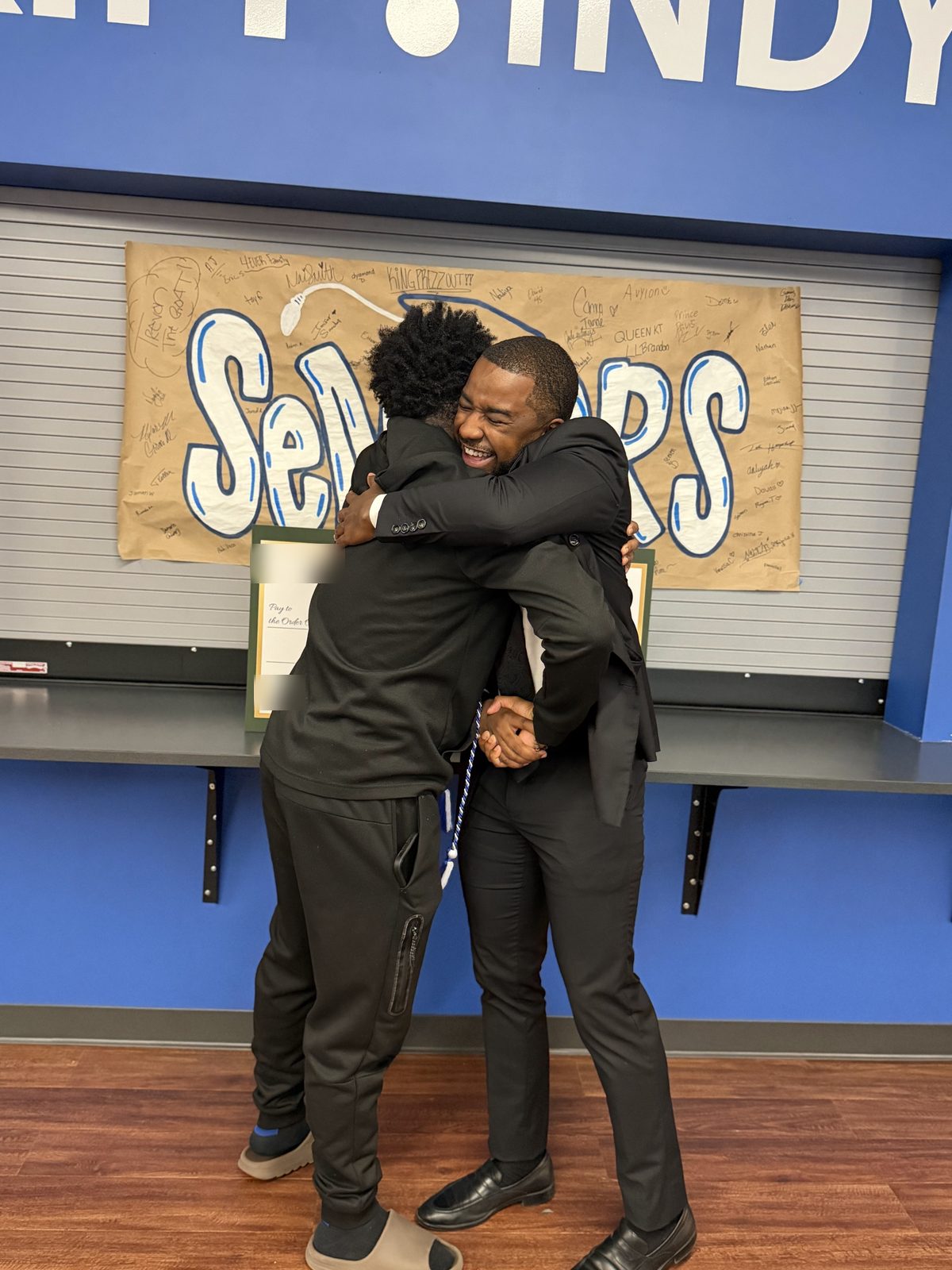 The graduate and presenter share a hug in front of the seniors banner