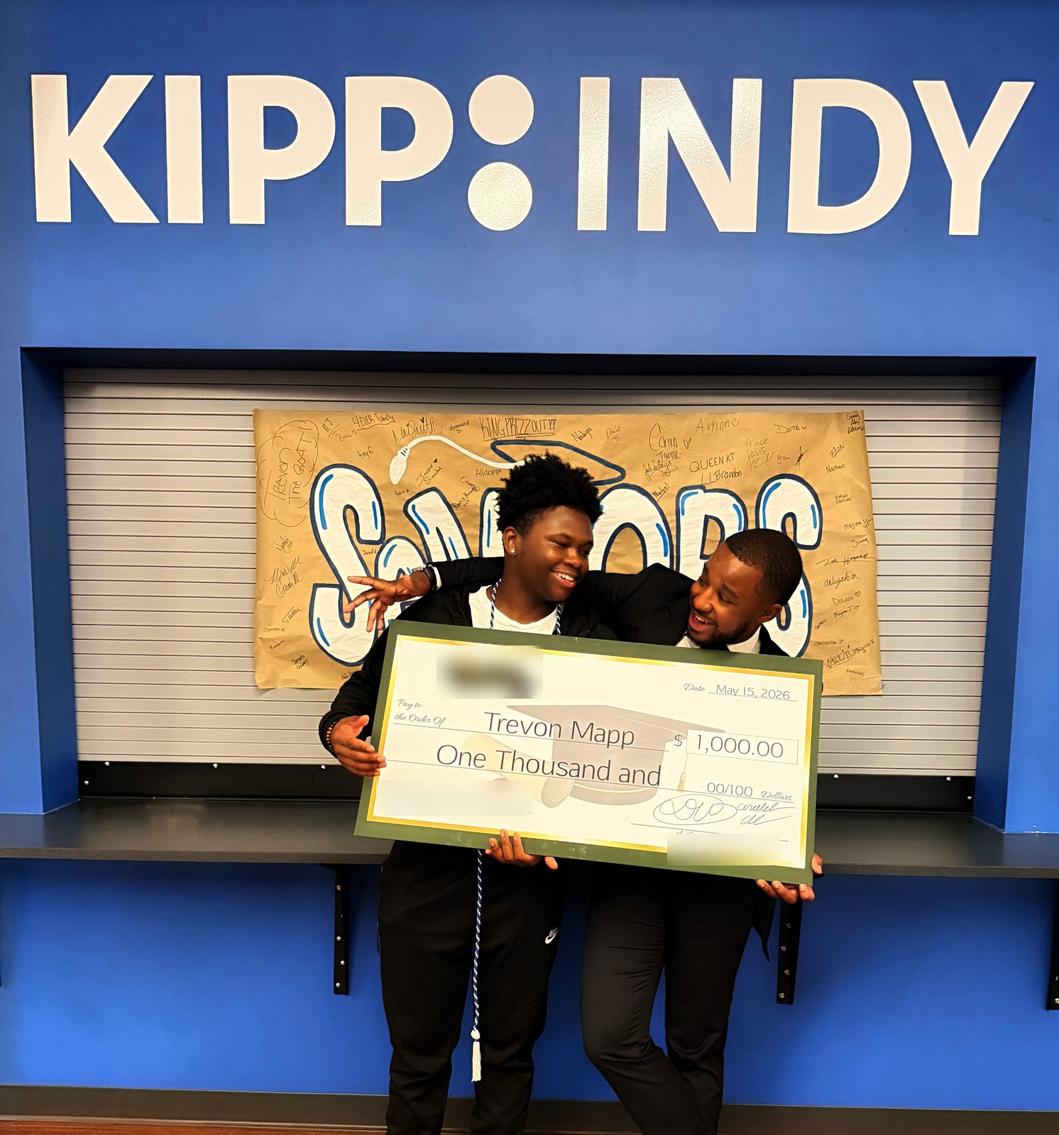 The graduate and presenter pose with the check under a KIPP Indy sign and a signed seniors banner