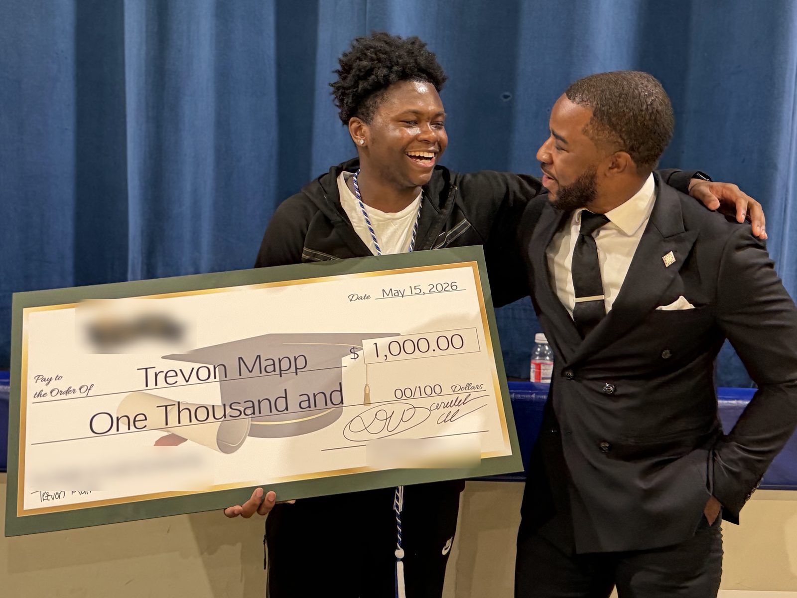 A graduate laughs while holding a large $1,000 scholarship check beside the presenter