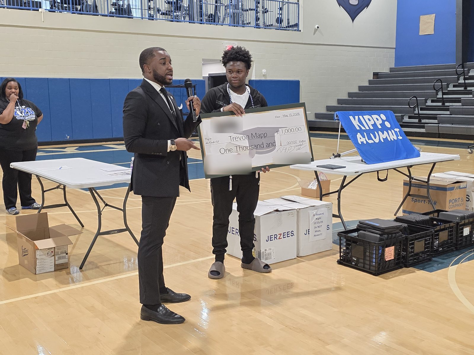 The presenter speaks into a microphone in a school gym while the graduate holds the scholarship check