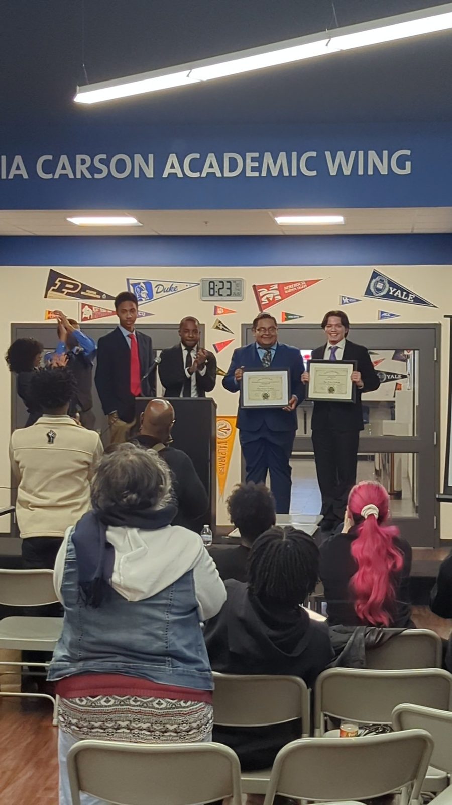 Two students hold framed certificates as others applaud on stage