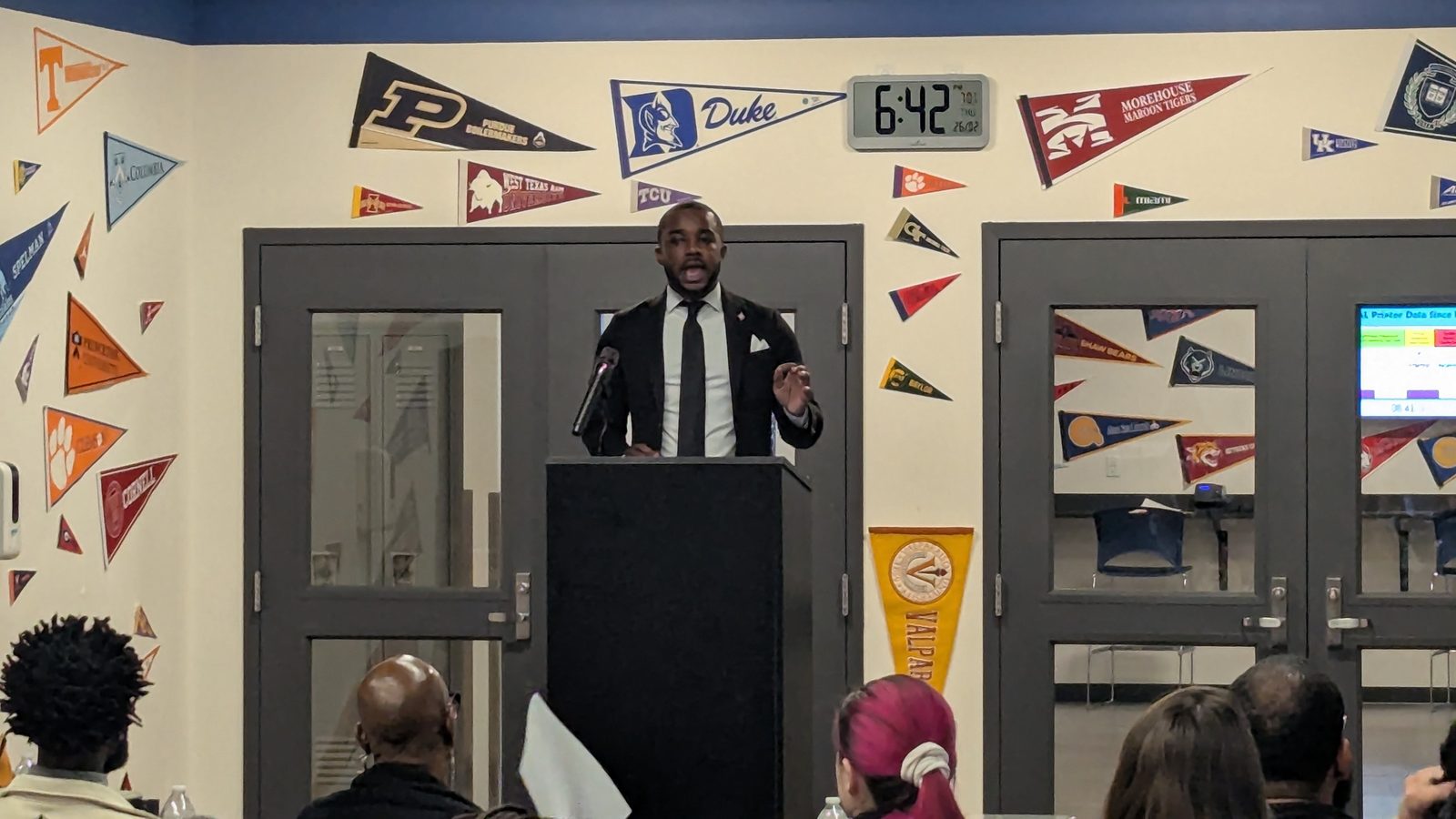 A speaker addresses the audience from the podium beneath a wall of college pennants