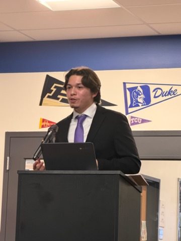 A student in a suit speaks at the podium in front of college pennants